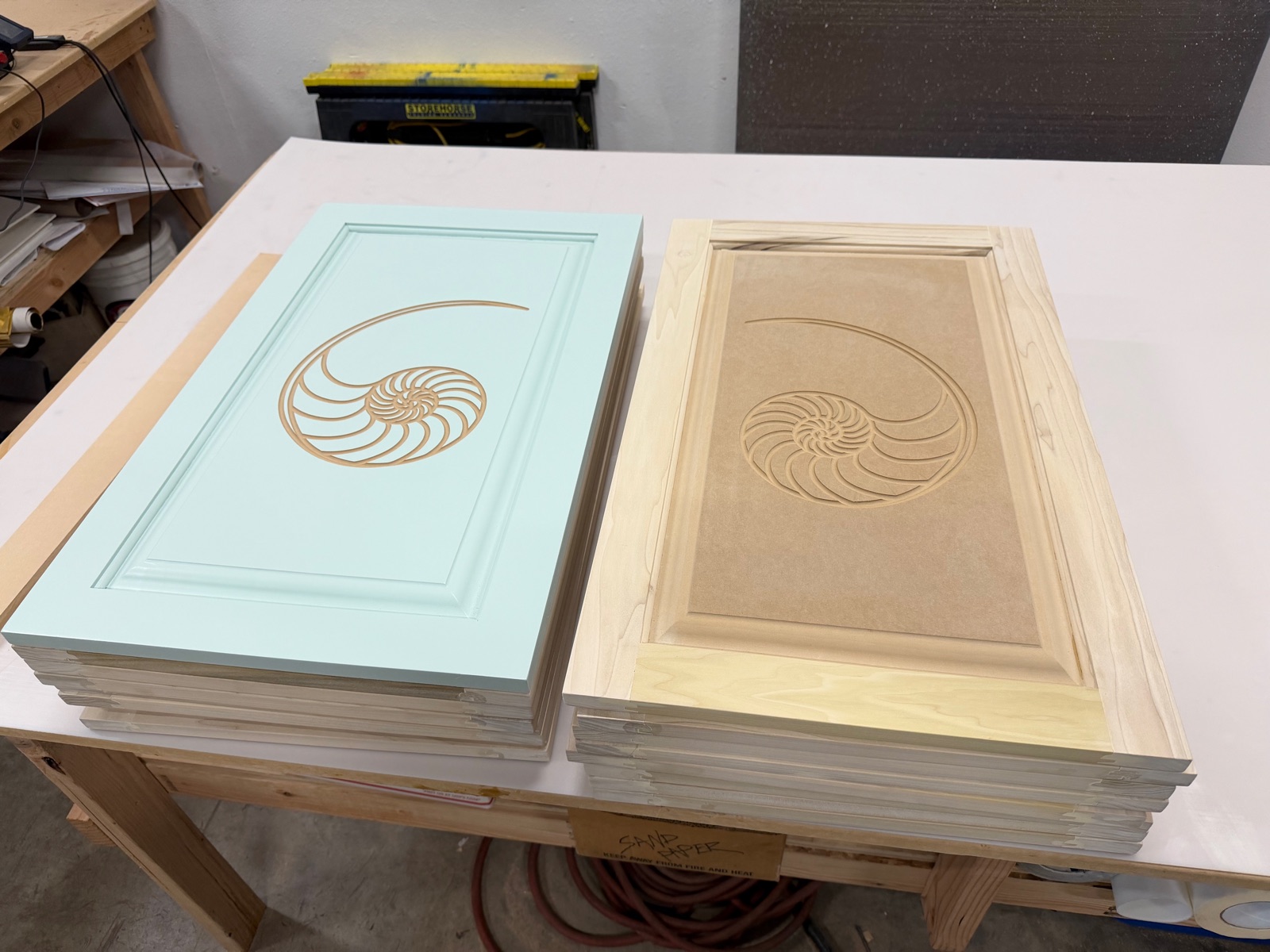 Stacks of engraved cabinet door faces from the production run, the client's mint finish sample beside a bare panel showing the mirrored nautilus
